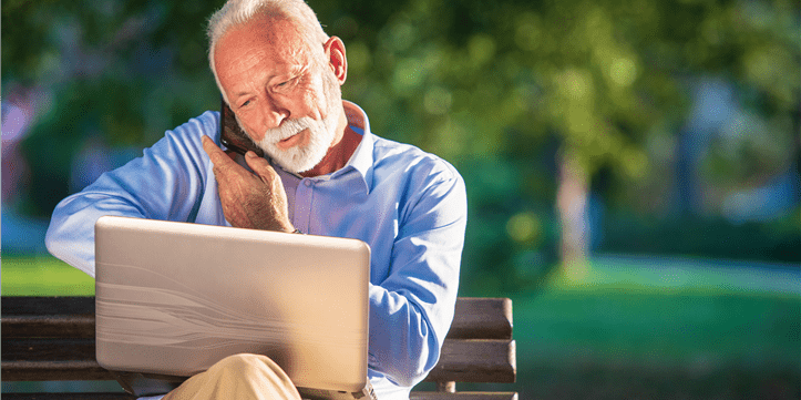 Vecchio signore seduto sulla panchina del parco, telefonando e avendo il computer portatile in ginocchio.