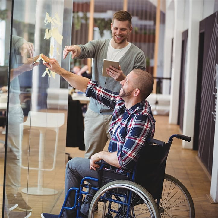 Ein Mann im Rollstuhl skizziert im Büro eine Idee mit seinem Arbeitskollegen an einer Glaswand.
