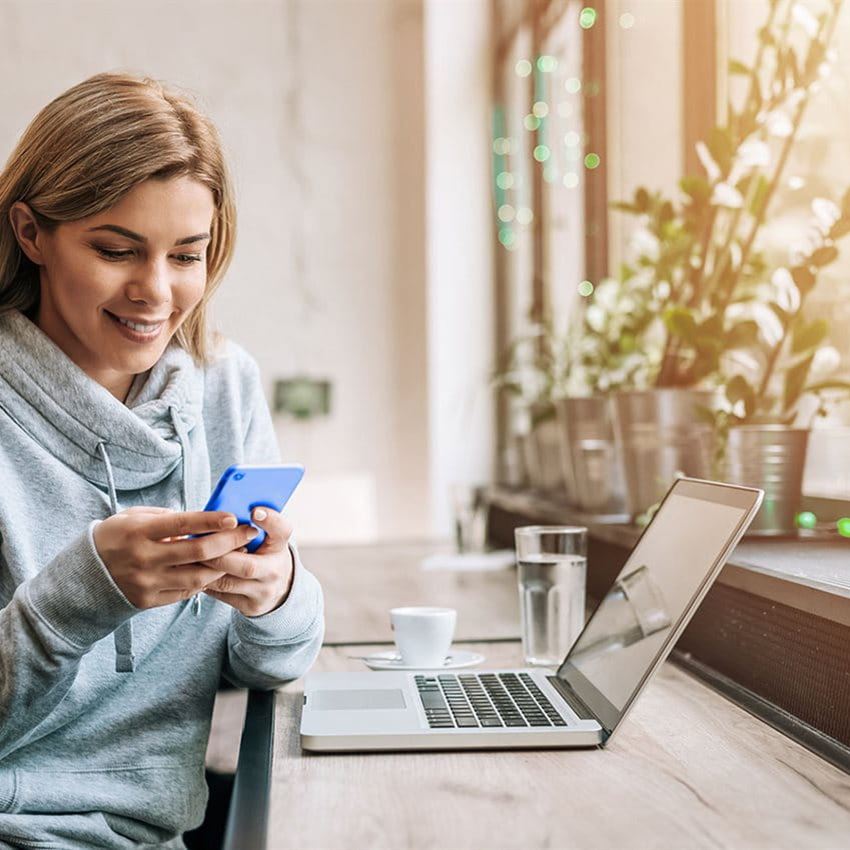 Eine junge Frau sitzt lächelnd an einem Fensterarbeitsplatz vor Ihrem Laptop und schaut auf ihr Mobile.