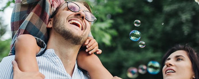 A child sitting on his father's shoulders grabs soap bubbles created by his mother.