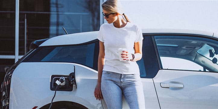 Frau steht vor ihrem Auto an der Elektroladestation und trinkt Kaffee