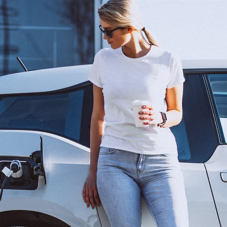 Une femme se tient devant sa voiture à la station de charge électrique et boit du café