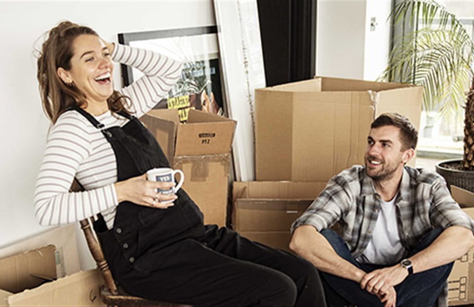 A couple is sitting between moving boxes and laughing.