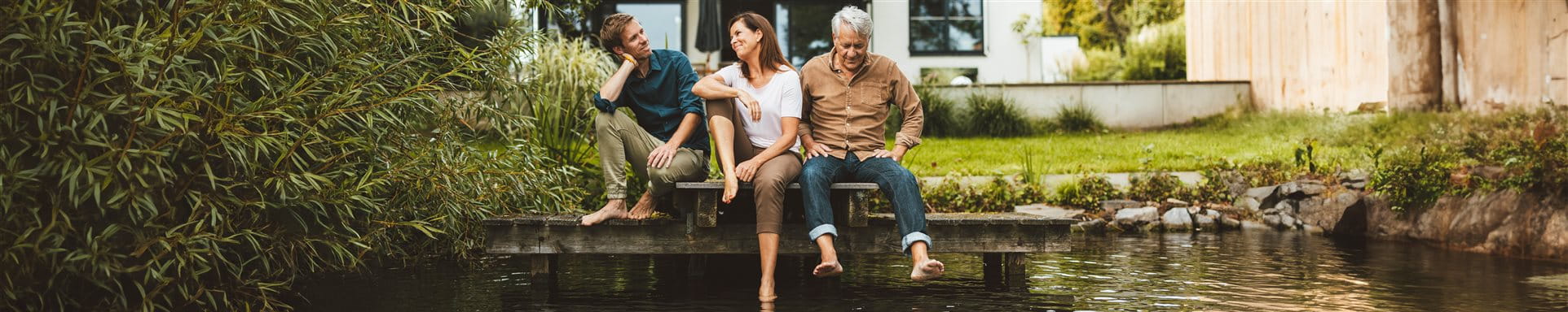 Drei Personen sitzen auf einem Steg an einem Teich vor dem Haus