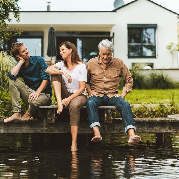 Drei Personen sitzen auf einem Steg an einem Teich vor dem Haus